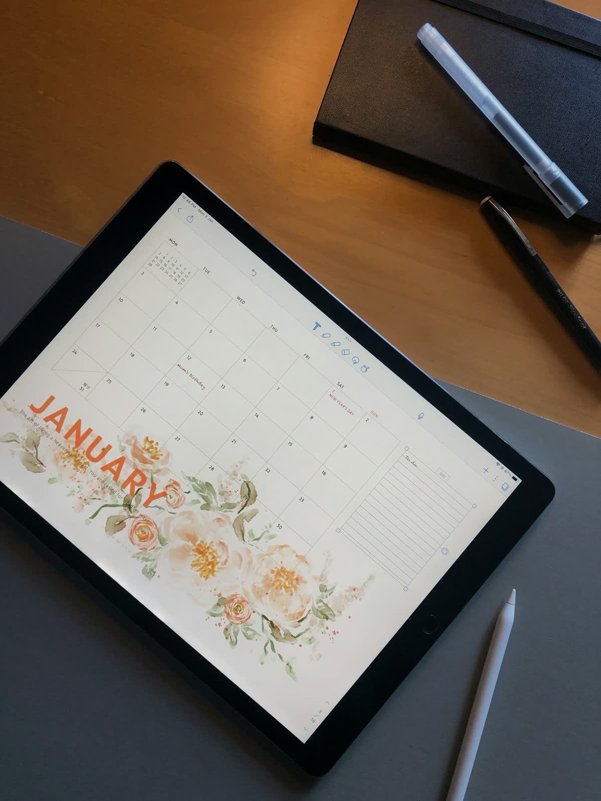 Tablet z kalendarzem miesięcznym leżący na biurku obok długopisu i notesu